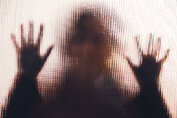 Back lit image of the silhouette of a woman with her hands pressed against a glass window. The silhouette is distorted, and the arms elongated, giving an alien-like quality. The image is sinister and foreboding, with an element of horror. It is as if the 'woman' is trying to escape from behind the glass.